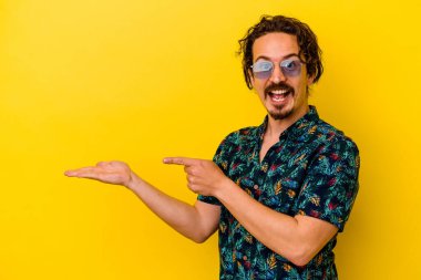 Sarı arka planda izole edilmiş yaz kıyafetleri giyen genç beyaz adam, palmiye ağacında bir kopya alanı tutarken heyecanlanıyor..