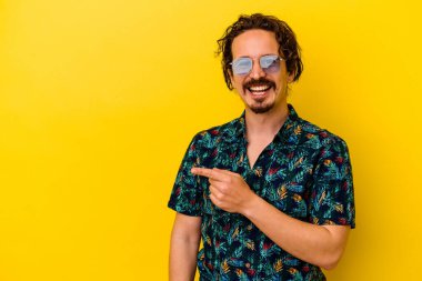 Sarı arka planda izole edilmiş yaz kıyafetleri giyen genç beyaz adam gülümsüyor ve boş alanda bir şeyler gösteriyor..