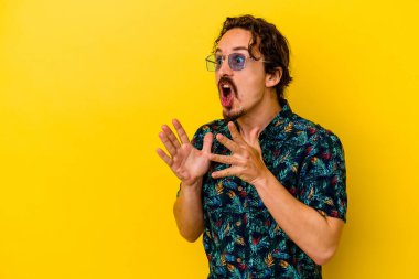 Sarı arka planda izole edilmiş yaz kıyafetleri giyen genç beyaz adam yüksek sesle bağırıyor, gözleri açık ve elleri gergin tutuyor..