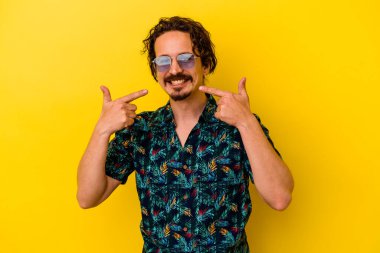 Sarı arka planda izole edilmiş yaz kıyafetleri giyen genç beyaz adam gülümsüyor ve parmaklarını ağzına doğrultuyor..