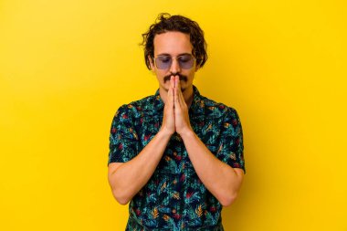Sarı arka planda izole edilmiş yaz kıyafetleri giyen genç beyaz bir adam el ele tutuşup ağız kenarında dua ediyor, kendinden emin hissediyor..