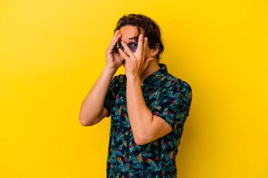 Sarı arka planda izole edilmiş yaz kıyafetleri giyen genç beyaz adam parmaklarının arasından korkmuş ve gergin bir şekilde göz kırpıyor..
