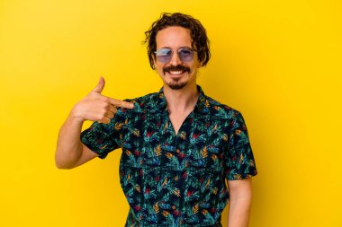 Sarı arka plandaki izole edilmiş yaz kıyafetleri giyen genç beyaz adam bir gömlek fotokopi alanını işaret ediyor, gururlu ve kendinden emin.