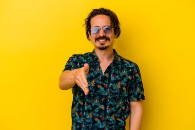 Sarı arka planda izole edilmiş yaz kıyafetleri giyen genç beyaz adam kameraya doğru elini uzatarak selam veriyor..