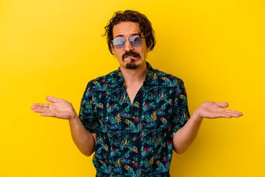 Sarı arka planda izole edilmiş yaz kıyafetleri giyen genç beyaz bir adam. Kopya alanını tutmak için omuz silkiyor ve kafası karışık..