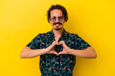 Sarı arka planda izole edilmiş yaz kıyafetleri giyen genç beyaz adam gülümsüyor ve elleriyle kalp şeklini gösteriyor..