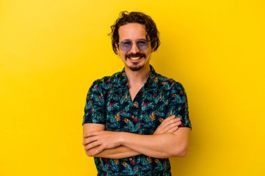 Sarı arka planda izole edilmiş yaz kıyafetleri giyen genç beyaz bir adam kendini güvende hissediyor ve kararlılıkla kollarını kavuşturuyor..