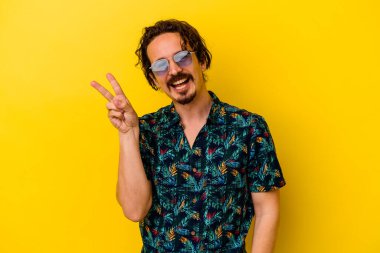 Sarı arka planda izole edilmiş yaz kıyafetleri giyen genç beyaz adam neşeli ve kaygısız parmaklarıyla barış sembolü gösteriyor..