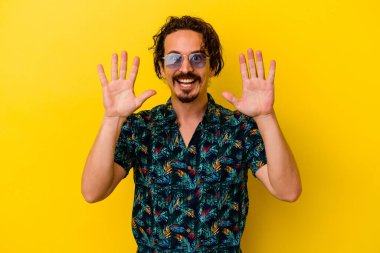 Sarı arka planda izole edilmiş yaz kıyafetleri giyen genç beyaz adam on numarayı elleriyle gösteriyor..