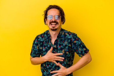 Sarı arka planda izole edilmiş yaz kıyafetleri giyen genç beyaz adam mutlu bir şekilde gülüyor ve ellerini karnının üstünde tutarak eğleniyor..