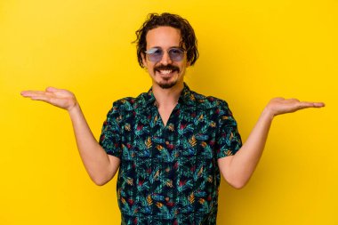 Sarı arka planda izole edilmiş yaz kıyafetleri giyen genç beyaz adam kollarıyla ölçek oluşturuyor, mutlu ve kendinden emin hissediyor..