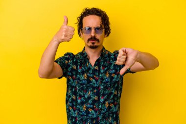 Sarı arka planda izole edilmiş yaz kıyafetleri giyen genç beyaz adam baş parmağını kaldırıyor ve baş parmak aşağı, zor seçim kavramı gösteriyor.