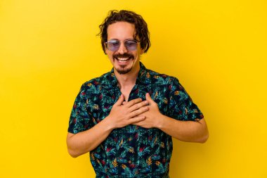 Sarı arka planda izole edilmiş yaz kıyafetleri giyen genç beyaz adam gülüyor, ellerini kalbinde tutuyor, mutluluk kavramını koruyor..