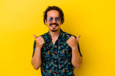Sarı arka planda izole edilmiş yaz kıyafetleri giyen genç beyaz adam iki parmağını da kaldırıyor, gülümsüyor ve kendinden emin..