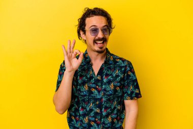 Sarı arka planda izole edilmiş yaz kıyafetleri giyen genç beyaz bir adam göz kırpıyor ve elleriyle iyi bir jest yapıyor..