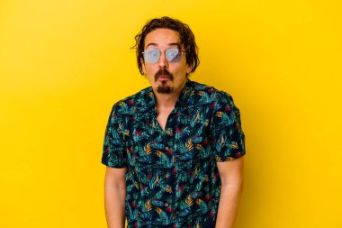 Sarı arka planda izole edilmiş yaz kıyafetleri giyen genç beyaz adam omuzlarını silker ve gözleri açık kalır..