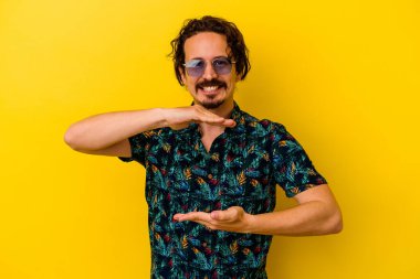 Sarı arka planda izole edilmiş yaz kıyafetleri giyen genç beyaz adam iki eliyle bir şey tutuyor, ürün sunumu..