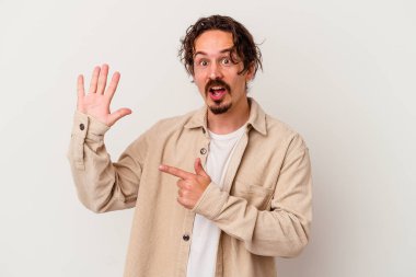 Beyaz arka planda izole edilmiş genç beyaz bir adam gülümseyerek beş numarayı parmaklarıyla gösteriyor..