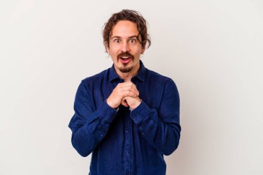 Beyaz arka planda izole edilmiş genç beyaz bir adam şans için dua ediyor, şaşırmış ve ağzını açıp önüne bakıyor..