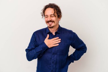 Beyaz arka planda soyutlanmış beyaz tenli genç bir adam gülüyor, elini yüreğine koyuyor, mutluluk kavramını koruyor..