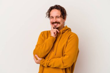 Beyaz arka planda izole olmuş genç beyaz bir adam gülümsüyor ve kendinden emin, çenesine elleriyle dokunuyor..