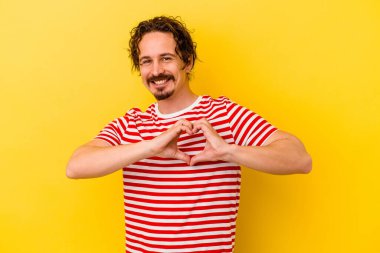 Sarı arka planda izole edilmiş genç beyaz adam gülümsüyor ve elleriyle kalp şeklini gösteriyor..