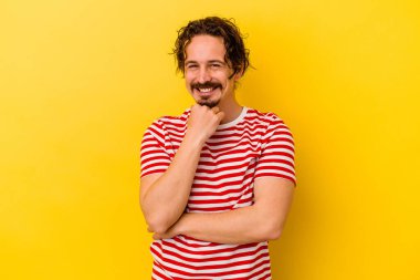 Sarı arka planda izole edilmiş genç, mutlu ve kendinden emin gülümseyen, elleriyle çenesine dokunan beyaz bir adam..