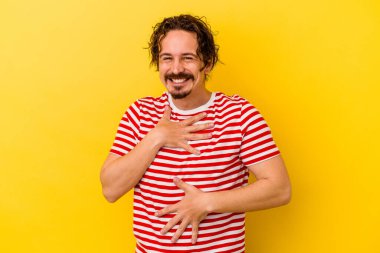 Sarı arka planda izole edilmiş genç beyaz adam mutlu bir şekilde gülüyor ve ellerini karnının üstünde tutarak eğleniyor..