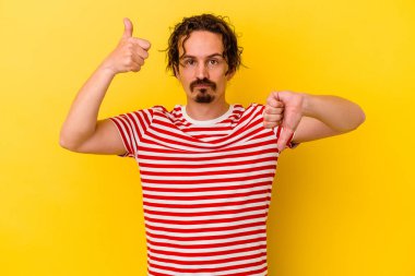 Sarı arka planda izole edilmiş genç beyaz adam baş parmağını kaldırıyor ve baş parmak aşağı, zor seçim konsepti.
