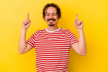 Sarı arka planda izole edilmiş genç beyaz bir adam iki parmağı da boş bir alan gösteriyor..