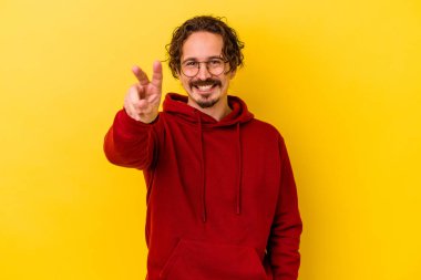 Sarı arka planda izole edilmiş genç beyaz adam zafer işareti gösteriyor ve gülümsüyor..