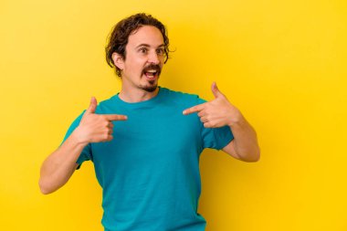 Sarı arka planda izole edilmiş genç beyaz adam parmağını göstererek şaşırdı ve gülümsedi..