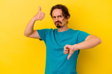 Sarı arka planda izole edilmiş genç beyaz adam baş parmağını kaldırıyor ve baş parmak aşağı, zor seçim konsepti.