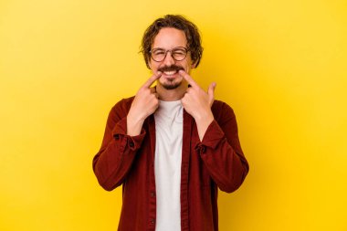 Sarı arka planda izole edilmiş genç beyaz adam gülümsüyor, parmaklarını ağzına doğrultuyor..