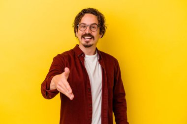 Sarı arka planda izole edilmiş genç beyaz adam selamlama jesti olarak kameraya elini uzatıyor..