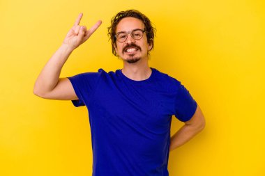 Sarı arka planda izole edilmiş genç beyaz adam parmaklarıyla kaya hareketi gösteriyor.