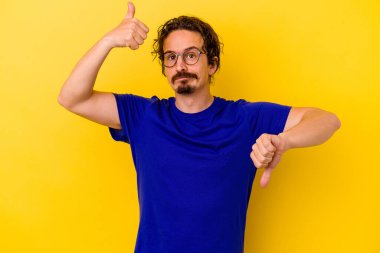 Sarı arka planda izole edilmiş genç beyaz adam baş parmağını kaldırıyor ve baş parmak aşağı, zor seçim konsepti.