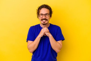 Sarı arka planda izole edilmiş genç beyaz adam ellerini çenesinin altında tutuyor ve mutlu bir şekilde yan tarafa bakıyor..