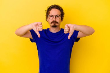 Sarı arka planda izole edilmiş genç beyaz adam baş parmağını aşağı gösteriyor, hayal kırıklığı konsepti..