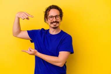 Sarı arka planda izole edilmiş genç beyaz adam işaret parmaklarıyla küçük bir şey tutuyor, gülümsüyor ve kendinden emin..