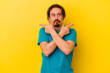 Sarı arka planda izole genç beyaz adam, iki seçenek arasında seçim yapmaya çalışıyor.