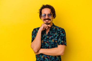 Sarı arka planda izole edilmiş yaz kıyafetleri giyen genç beyaz adam mutlu ve kendinden emin gülümsüyor, çenesine elleriyle dokunuyor..