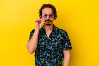 Sarı arka planda izole edilmiş yaz kıyafetleri giyen genç beyaz adam dudaklarında parmaklarıyla bir sır saklıyor..