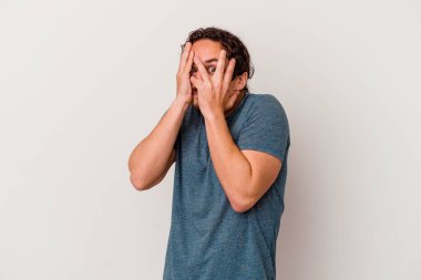 Beyaz arka planda izole edilmiş genç beyaz bir adam parmaklarının arasından korkmuş ve gergin bir şekilde göz kırpıyor..