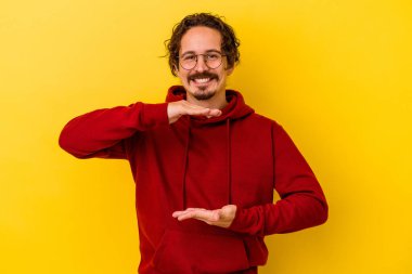 Sarı arka planda izole edilmiş genç beyaz adam iki eliyle bir şey tutuyor, ürün sunumu..