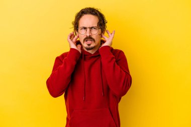 Sarı arka planda izole edilmiş genç beyaz bir adam parmaklarıyla kulaklarını kapatıyor, gürültülü bir ortam yüzünden stresli ve çaresiz..