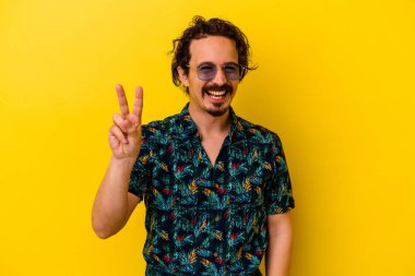 Sarı arka planda izole edilmiş yaz kıyafetleri giyen genç beyaz adam zafer işareti gösteriyor ve gülümsüyor..