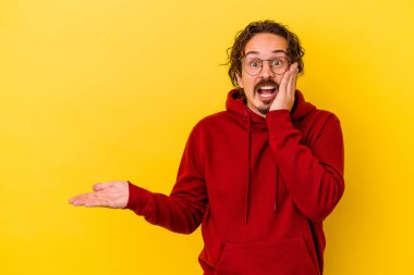 Sarı arka planda izole edilmiş genç beyaz adam, avuç içi fotokopi alanını elinde tutmaktan etkilendi..