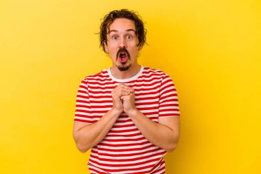 Kafkasyalı genç adam sarı arka planda izole edilmiş şans için dua ediyor, şaşırmış ve ağzını açıp önüne bakıyor..