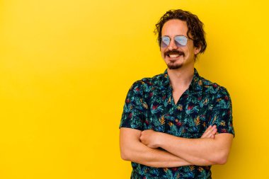 Sarı arka planda izole edilmiş yaz kıyafetleri giyen genç beyaz adam kollarını kavuşturarak gülümsüyor..